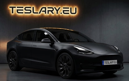 A matte black Tesla Model Y is shown from a three-quarter front angle in a dark studio setting. The background features a dark gray textured wall with a glowing orange 'TESLARY. Tesla Model 3 2019 2020 2021 2022 2023 Ubertine Wheel Cover Hubcaps 18inch Buy from Ireland and Europes Teslary.eu with Free Delivery with Fedex across Europe
