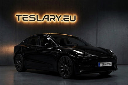 A black Tesla Model Y sedan shown from a three-quarter front angle in a dark studio setting, with a glowing 'TESLARY.EU' sign on the wall behind it and a matching license plate. Tesla_Model_3_2019_2020_2021_2022_2023_Ubertine_Wheel_Cover_Hubcaps_18inch_Buy_from_Ireland_and_Europes_Teslary.eu_with_Free_Delivery_with_Fedex_across_Europe_1_c60e6284-6ef1-49c7-aeb2-203f16d891